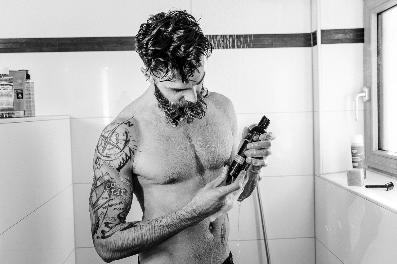 Mann hält unter der Dusche ein Bartshampoo in der Hand.