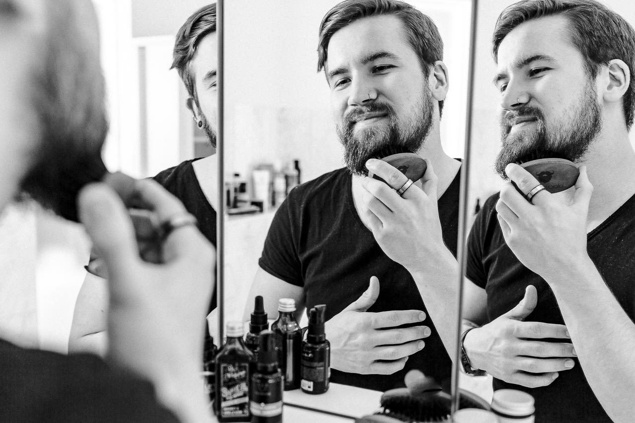 Mann bürstet seinen Bart vor dem Spiegel.